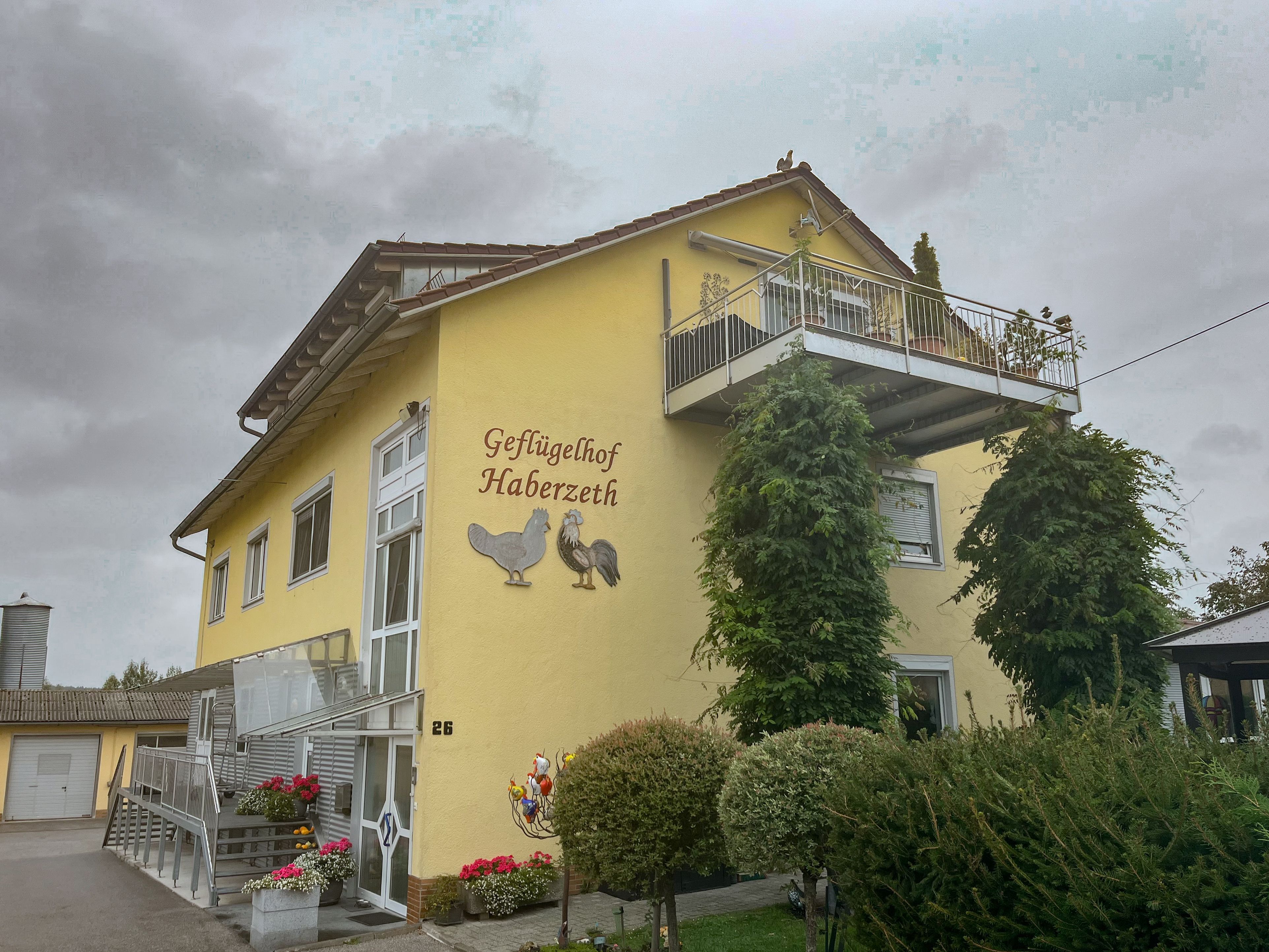 Gelb gestrichenes Wohn- und Betriebsgebäude des Geflügelhofs Haberzeth mit Balkon, begrünten Fassadenelementen und bepflanztem Eingangsbereich. An der Fassade ist die Aufschrift ‚Geflügelhof Haberzeth‘ mit Huhn- und Hahnmotiv angebracht.