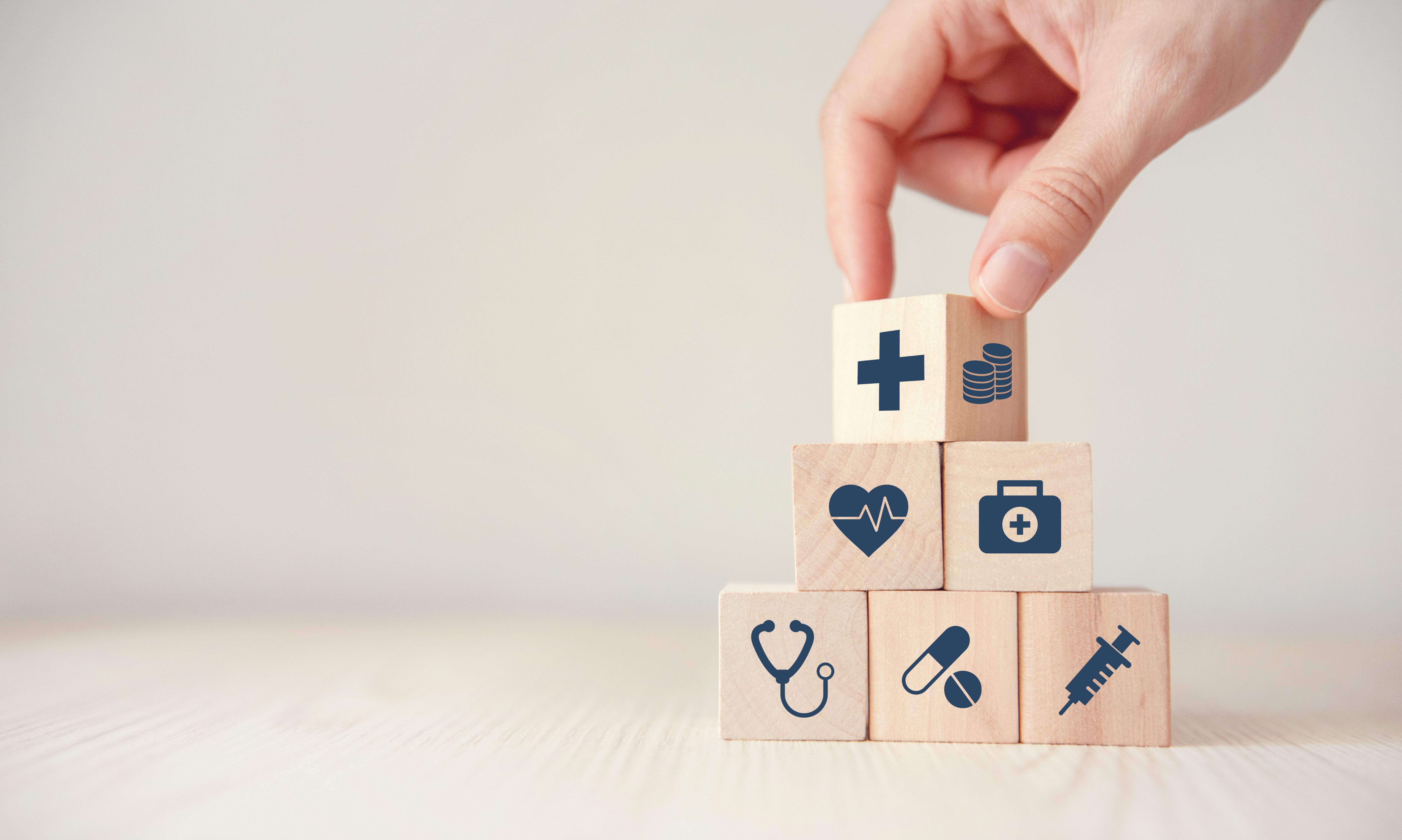 Hand stapelt kleine Holzwürfel zu einer Pyramide. Auf den Holzwürfeln sind Gesundheitssymbole, wie eine Spritze oder ein Stethoskope, zu sehen.