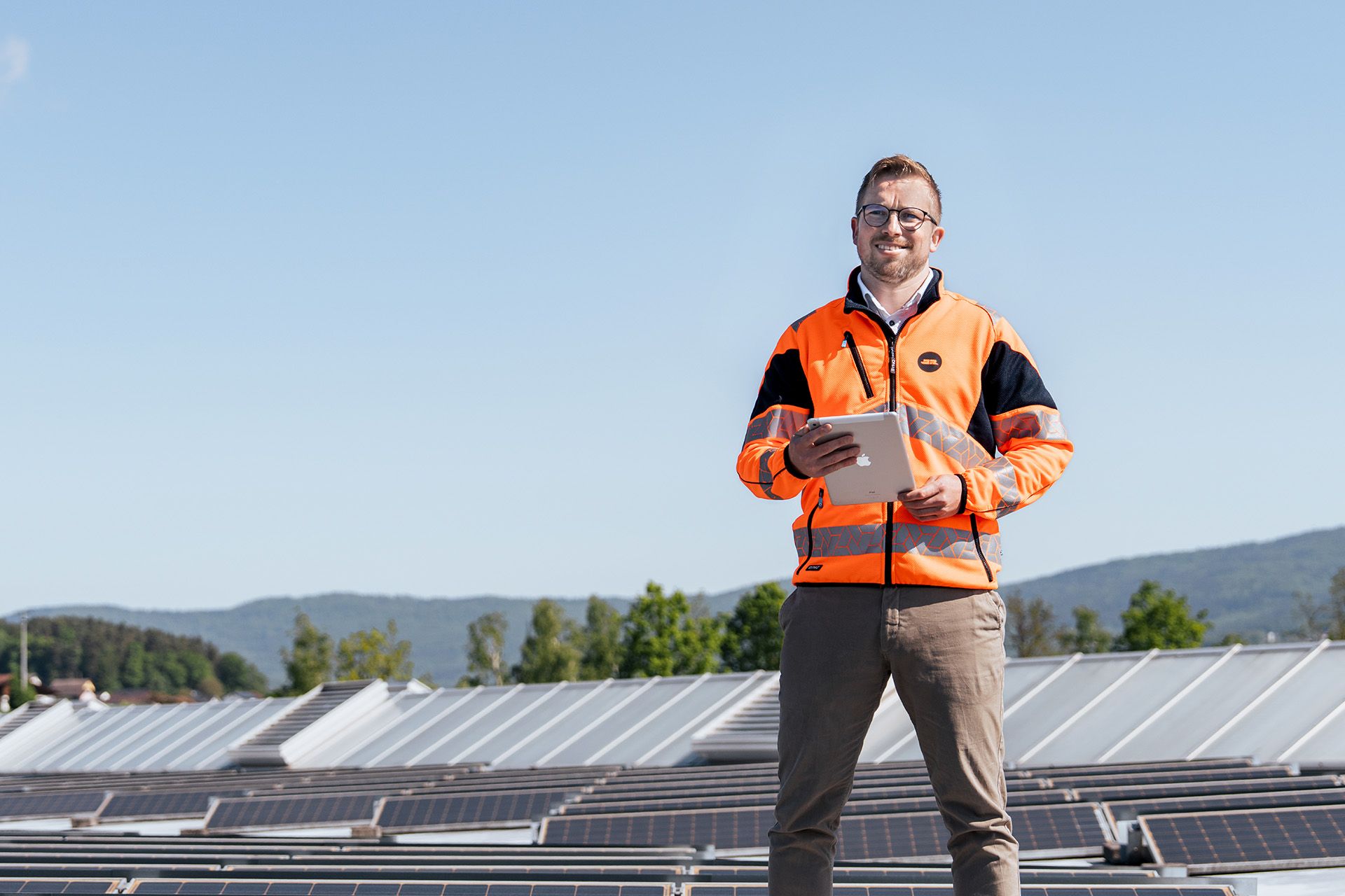 Energie- und Energiemanagement-Beauftragter Martin Wagner steht auf einem Dach mit Photovoltaikpaneelen, hält ein Tablet und trägt eine orange Sicherheitsjacke
