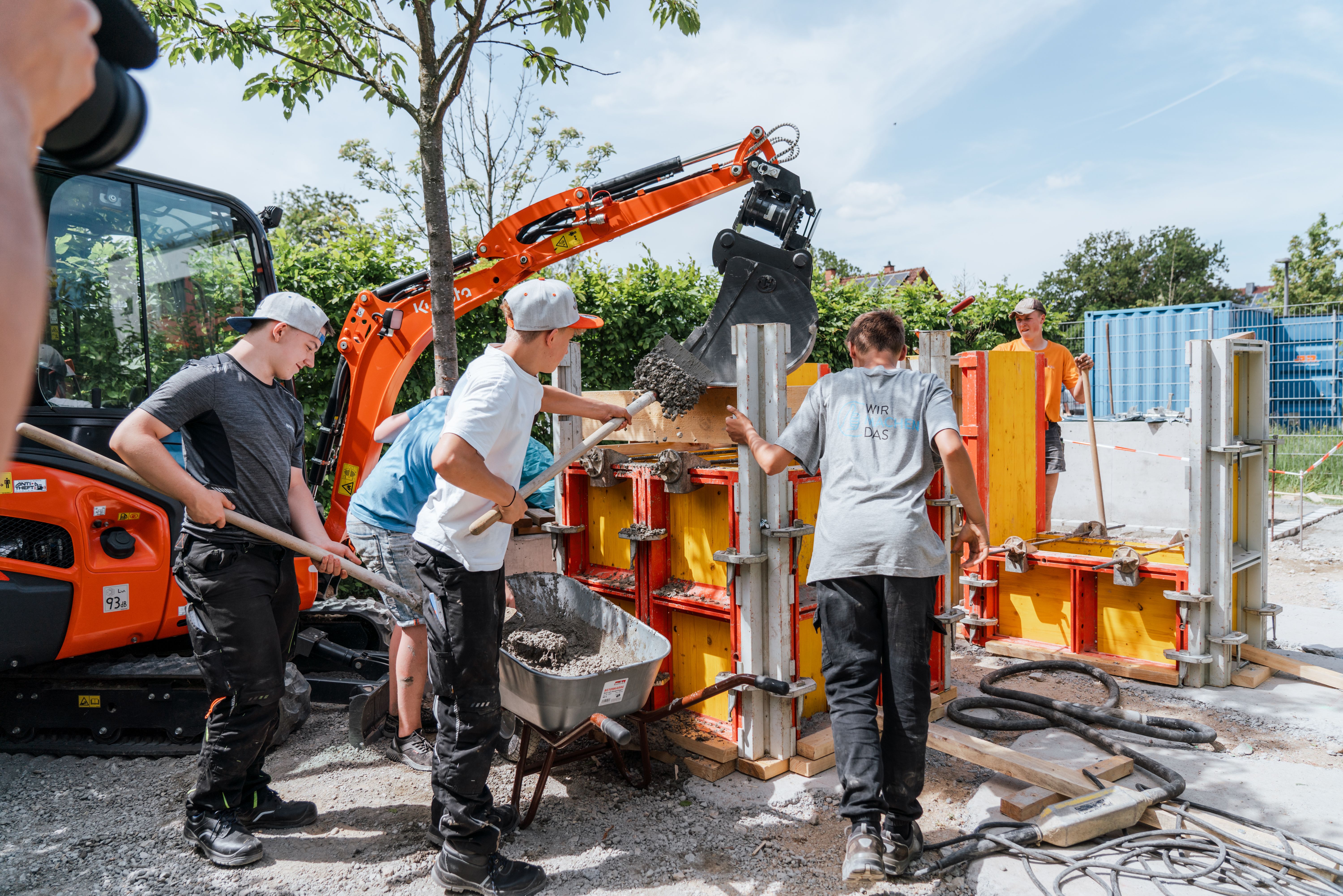 Der Beton wird mit Schaufeln in die Schalungen gefüllt. Ein Bagger unterstützt bei der Arbeit.