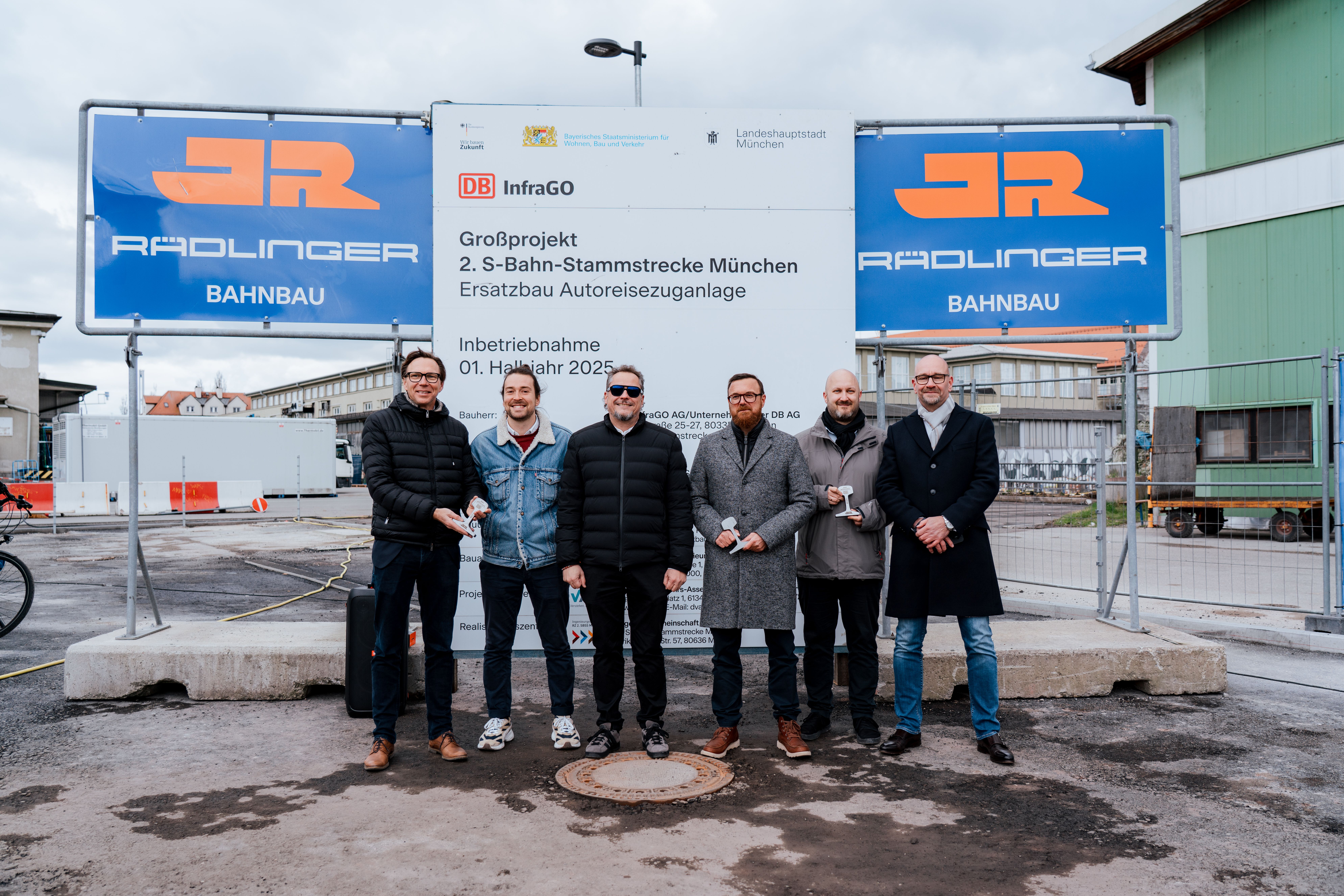 Gruppenfoto bei der Inbetriebnahme der Autoreisezuganlage München Süd: sechs Personen vor einem großen Schild, das auf das Großprojekt 2. S-Bahn-Stammstrecke München und den Ersatzbau der Autoreisezuganlage München Süd hinweist, links und rechts davon jeweils eine Bautafel mit dem Logo der Josef Rädlinger Unternehmensgruppe (Sparte Bahnbau)