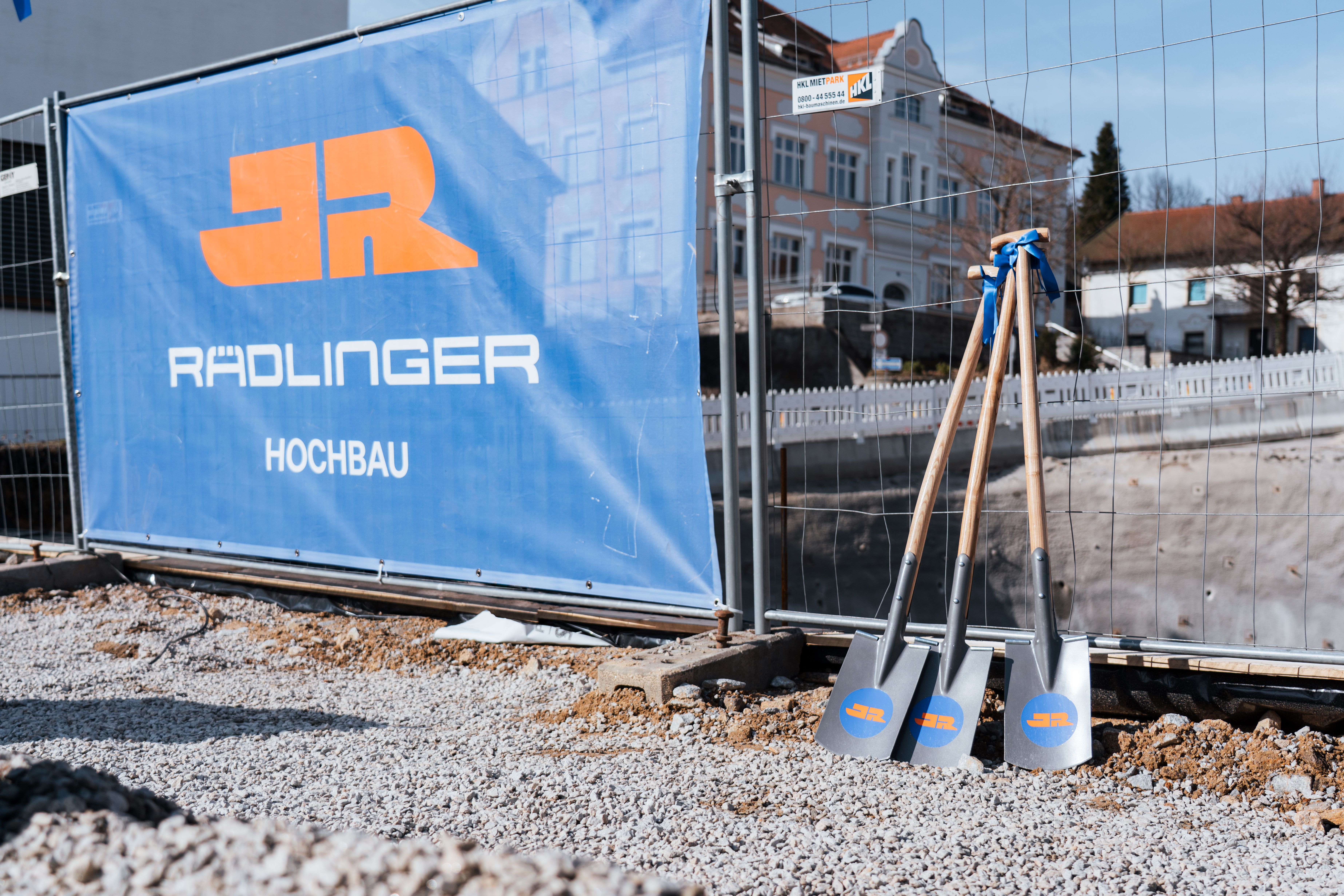 Drei Spaten lehnen am Bauzaun vor der Baugrube, daneben ein JR Banner