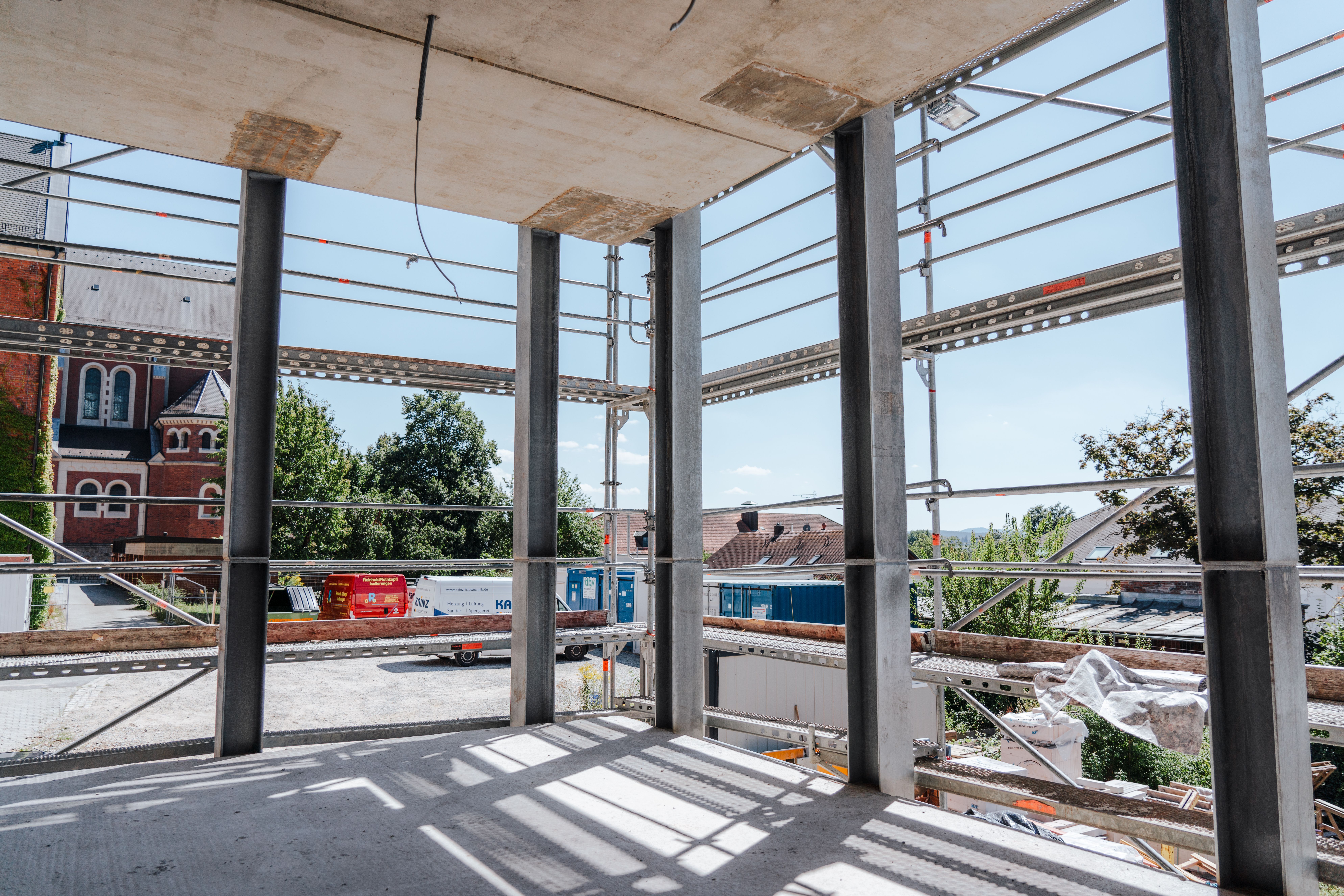 Blick aus dem Rohbau mit Stahlstützen und Betondecke. Das Gebäude ist von einem Gerüst umgeben. Im Hintergrund sind eine Kirche, Bäume und weitere Gebäude zu sehen. Auf dem Baugelände stehen mehrere Fahrzeuge und Container.