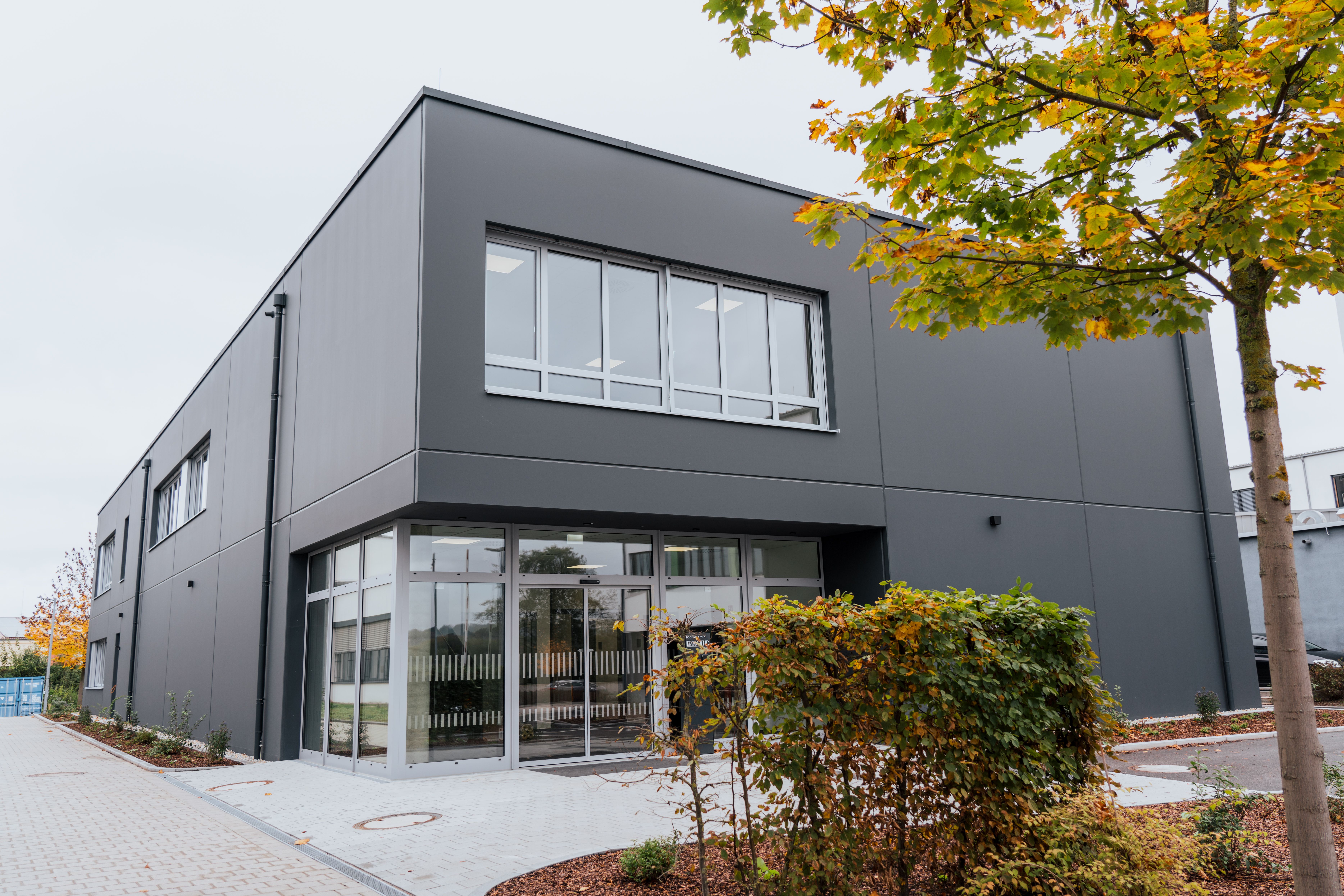 Das neue Campusgebäude von außen mit Blick auf den Haupteingang. Im Vordergrund einige Pflanzen und ein Baum mit Herbstlaub.