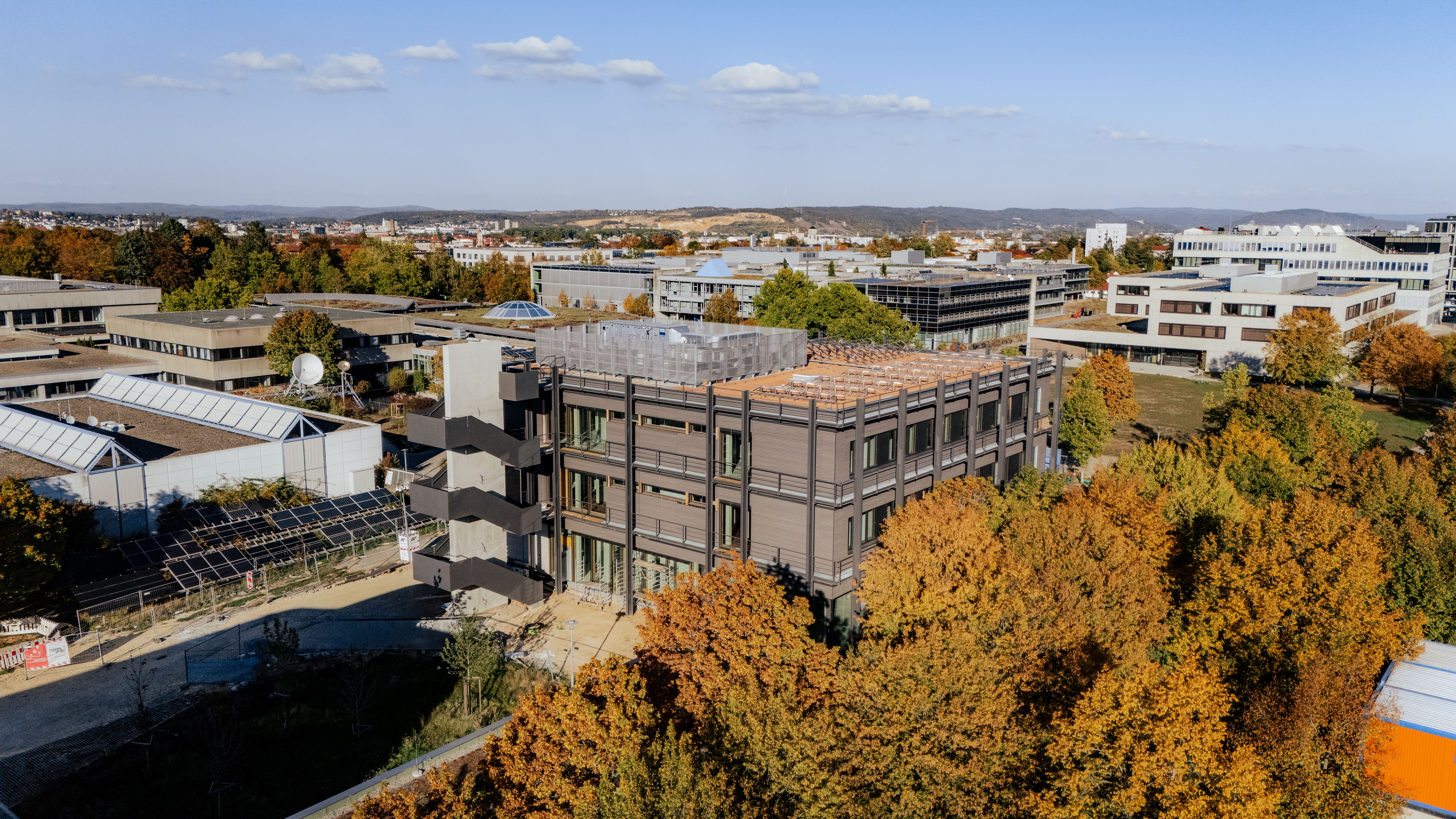 Luftaufnahme eines mehrgeschossigen Labor- und Bürogebäudes im Bau. Das Gebäude befindet sich auf einem umzäunten Baustellengelände und ist von weiteren Gebäuden sowie herbstlich gefärbten Bäumen umgeben.