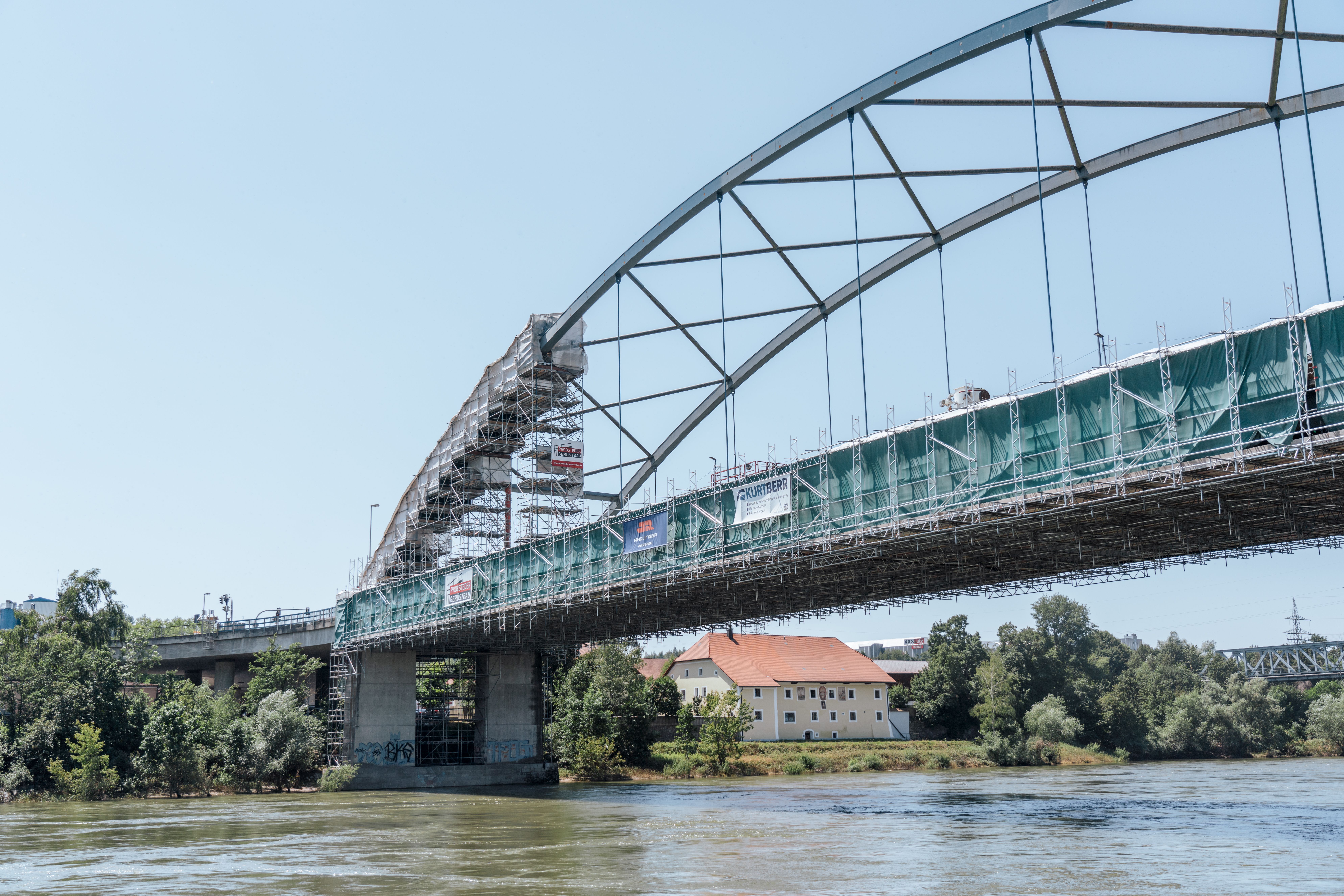 Eine große Stahlbogenbrücke über einen Fluss ist von einem umfangreichen Gerüst umgeben, das mit grünen Schutzplanen verkleidet ist. Mehrere Bauunternehmen haben ihre Banner am Gerüst angebracht. Im Hintergrund sind ein Gebäude mit rotem Dach und Bäume zu sehen. Der Himmel ist klar und blau.