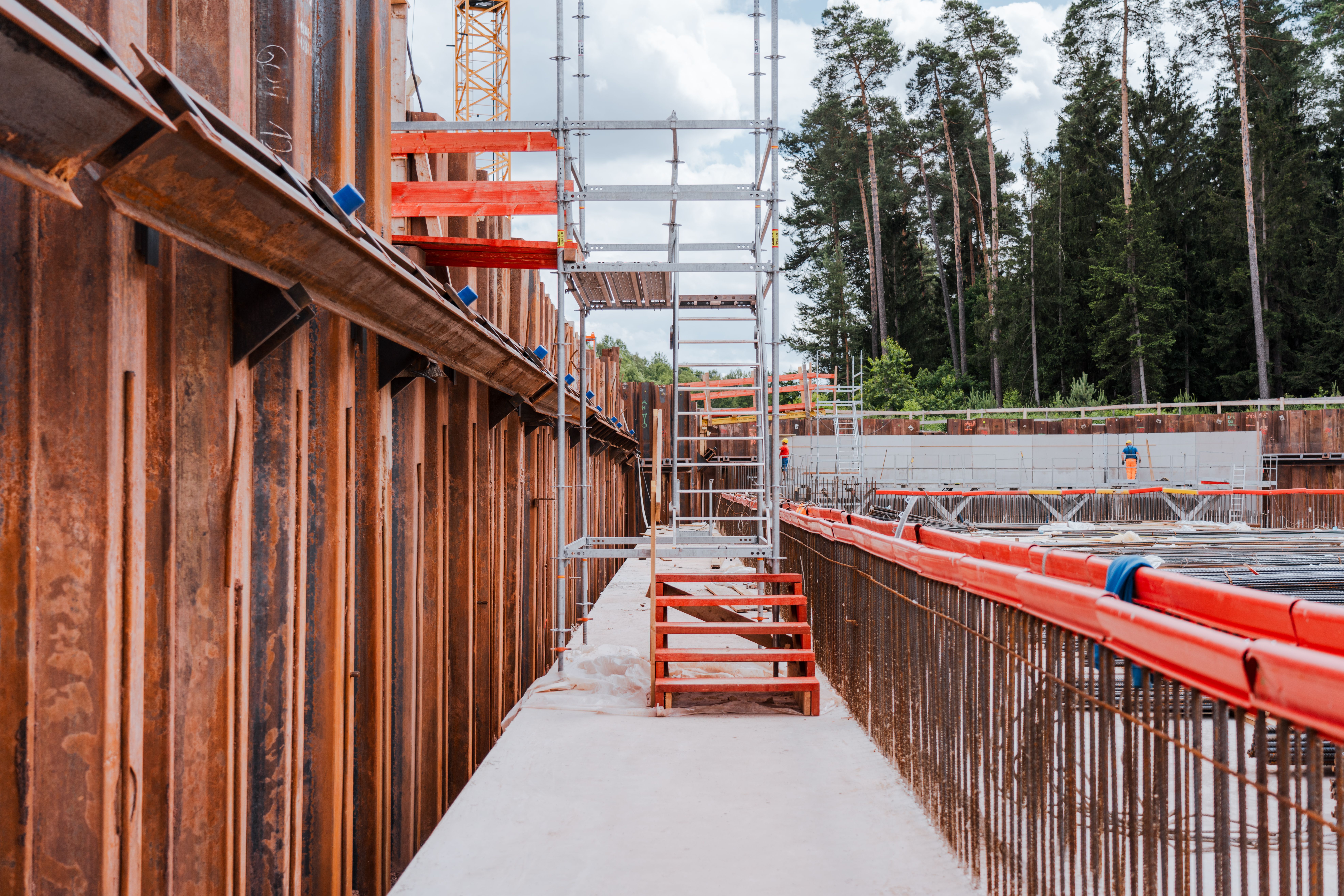 Schmale Betonlaufwege mit rotem Treppenzugang führen durch die Baustelle mit Stahlbewehrungen und einem Gerüst. Links ragen Spundwände empor, während im Hintergrund Bauarbeiter an einer Betonwand arbeiten. Dahinter erstreckt sich ein bewaldetes Gebiet.