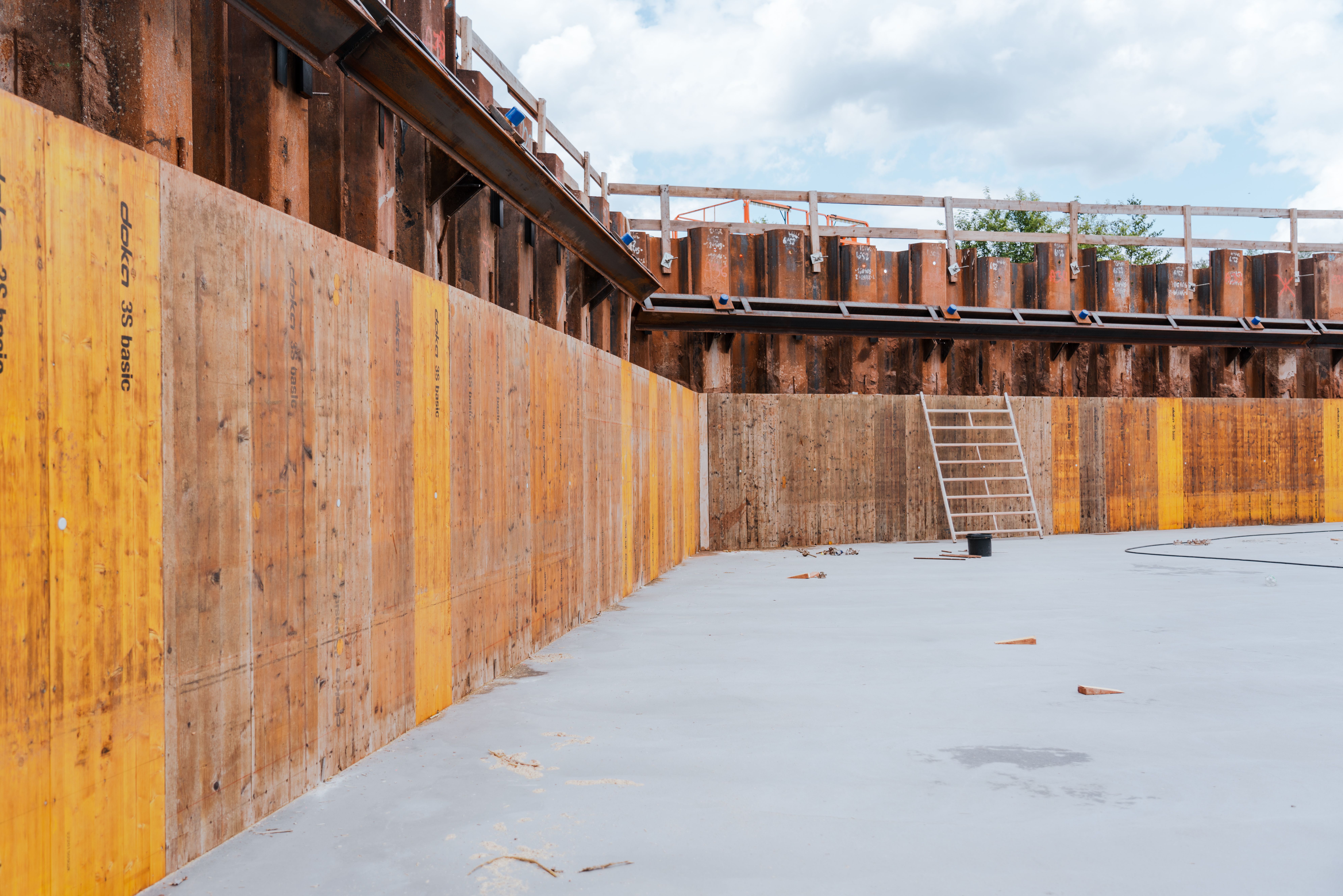 Betonierte Bodenfläche und Holzverschalungen. Spundwände sichern die Baugrube, und eine Leiter lehnt an der Wand für den Zugang zur oberen Ebene.
