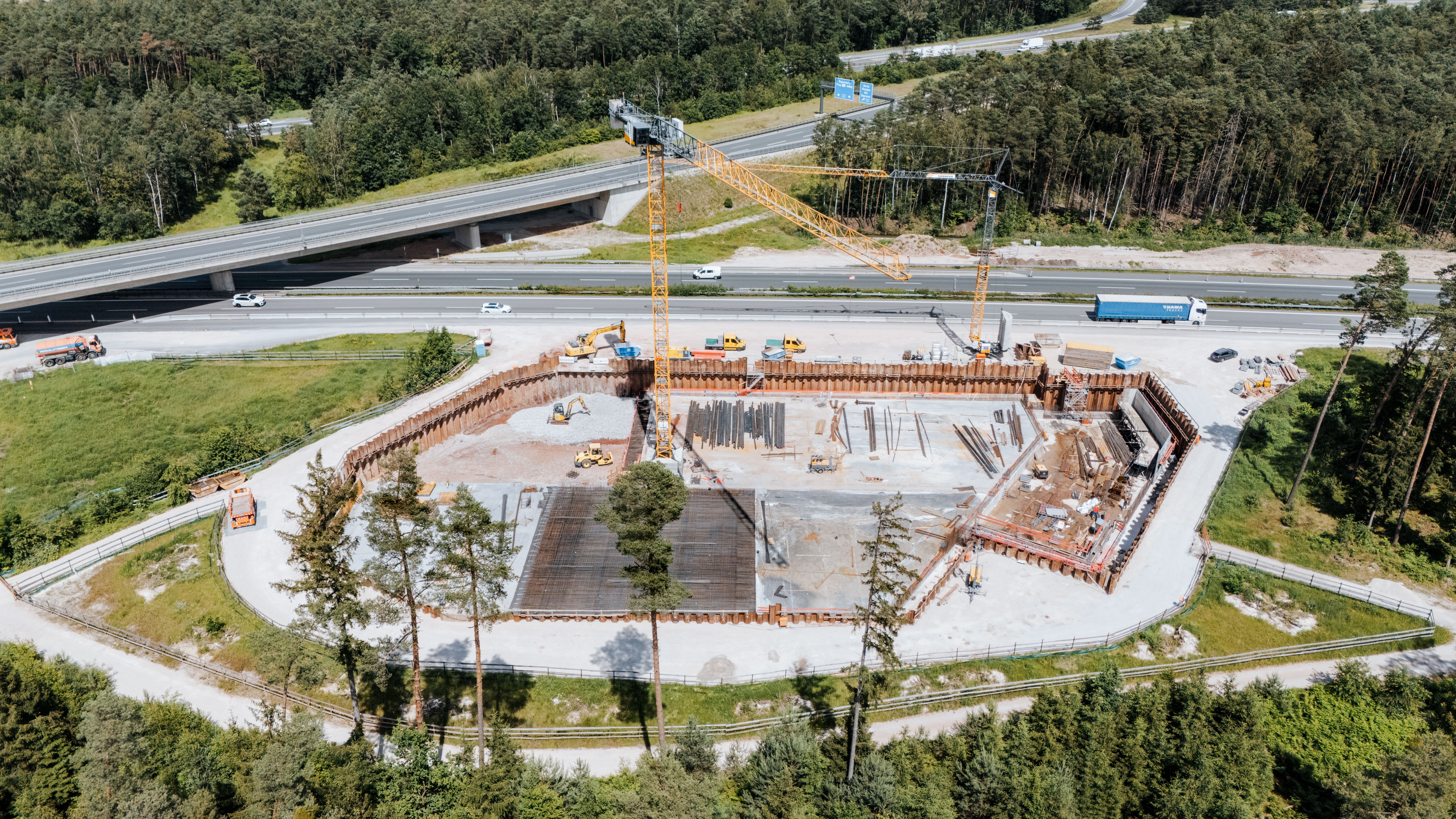 Luftaufnahme der Baustelle für das Regenrückhaltebecken neben der Autobahn. Die Baugrube ist mit Spundwänden gesichert, zwei Kräne stehen auf der Baustelle. Mehrere Baufahrzeuge und Baumaterialien sind auf dem Gelände verteilt. Im Hintergrund verläuft die Autobahn mit fahrenden Fahrzeugen, umgeben von Wald.