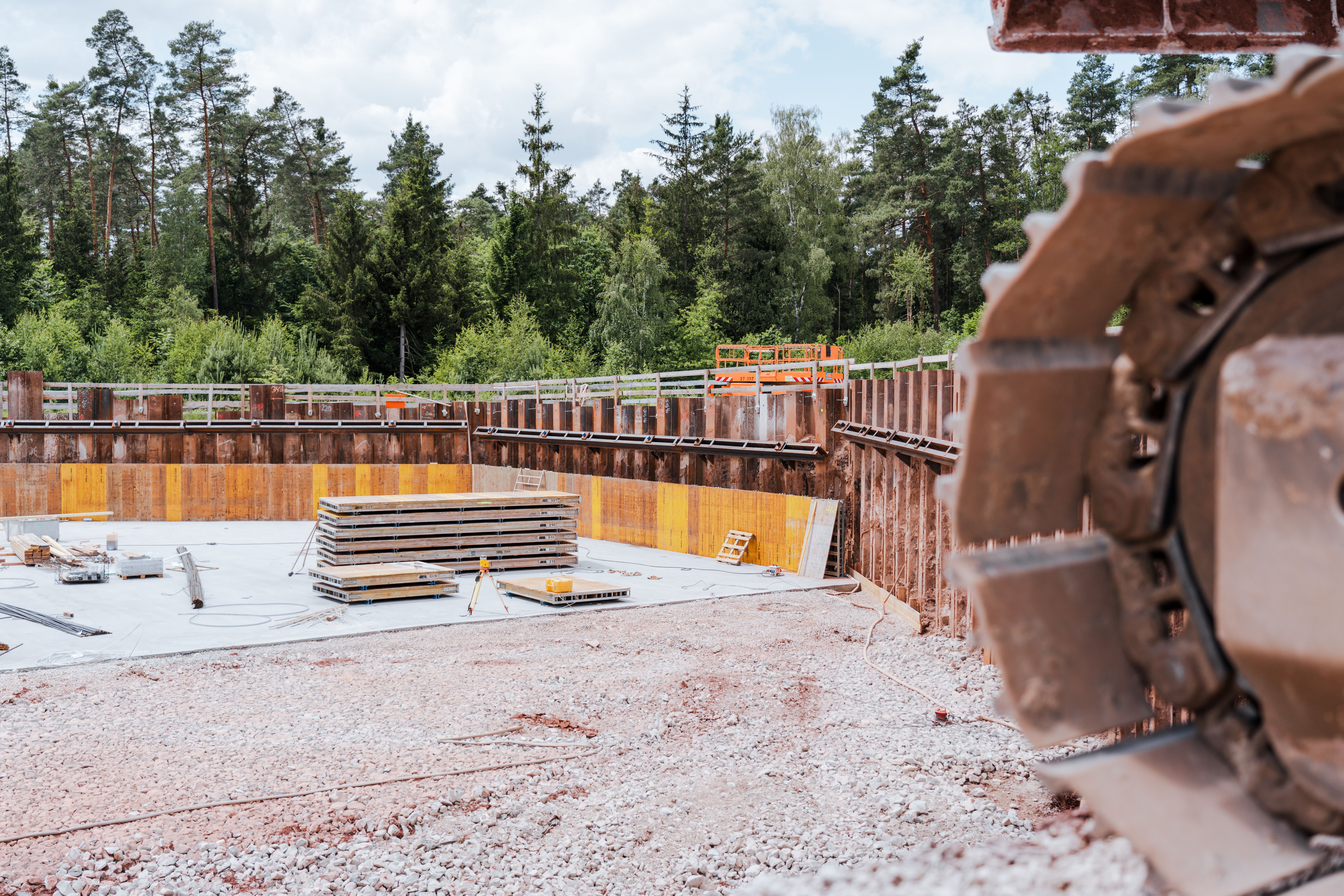 Ein großes, rundes Bauwerk als Begrenzung des Rückhaltebeckens mit Spundwänden und einer betonierten Bodenplatte ist zu sehen. In der Mitte liegen gestapelte Schalungselemente, während Vermessungsgeräte und Baumaterialien verteilt sind. Rechts im Vordergrund ist ein Teil eines schweren Bohrwerkzeugs zu erkennen. Im Hintergrund grenzt dichter Wald an die Baustelle.