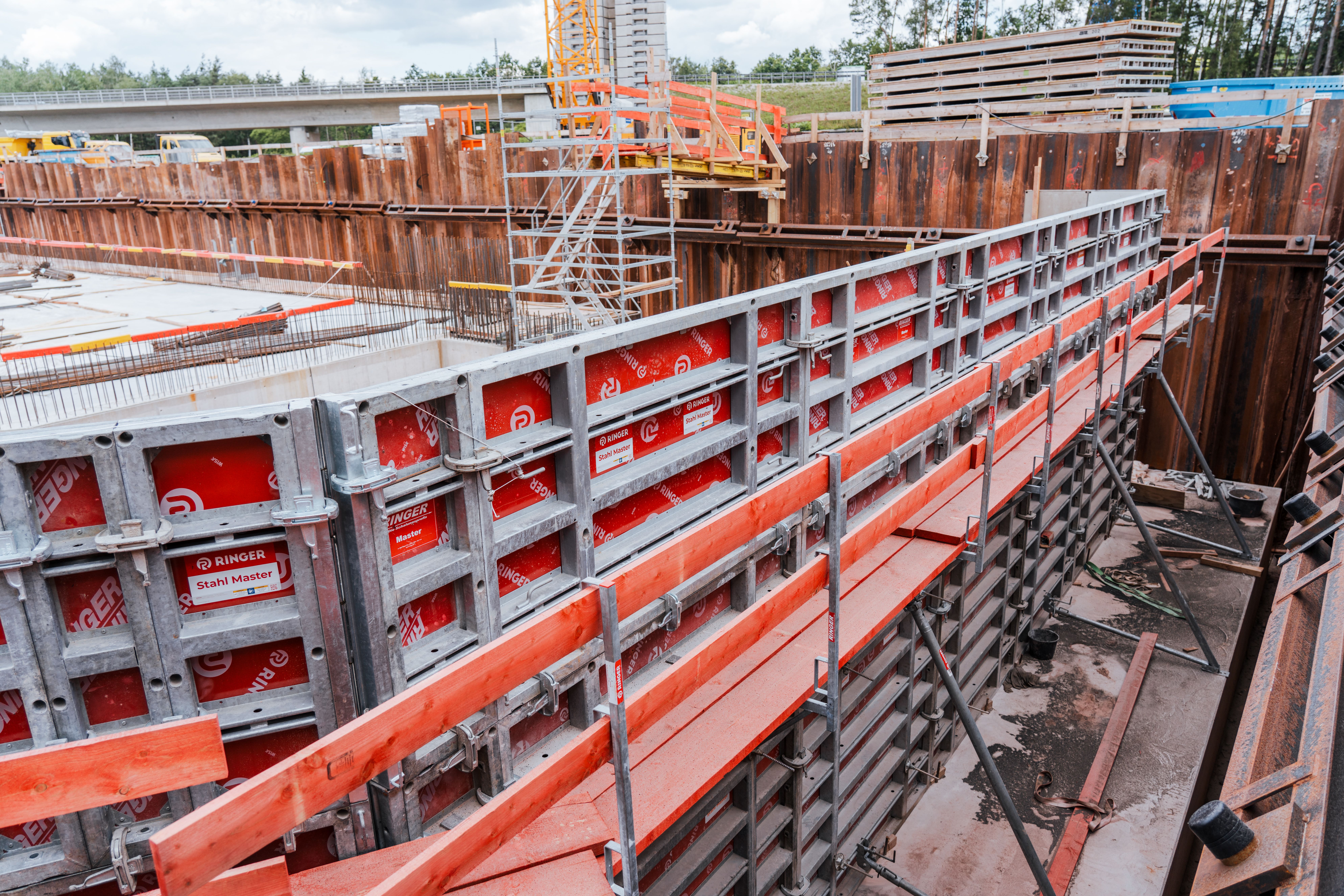 Zu sehen sind Stahlbeton-Schalungen. Rote Absturzsicherungen sind an den Schalungselementen befestigt. Im Hintergrund sind Spundwände, eine Treppe sowie eine Brücke zu sehen.