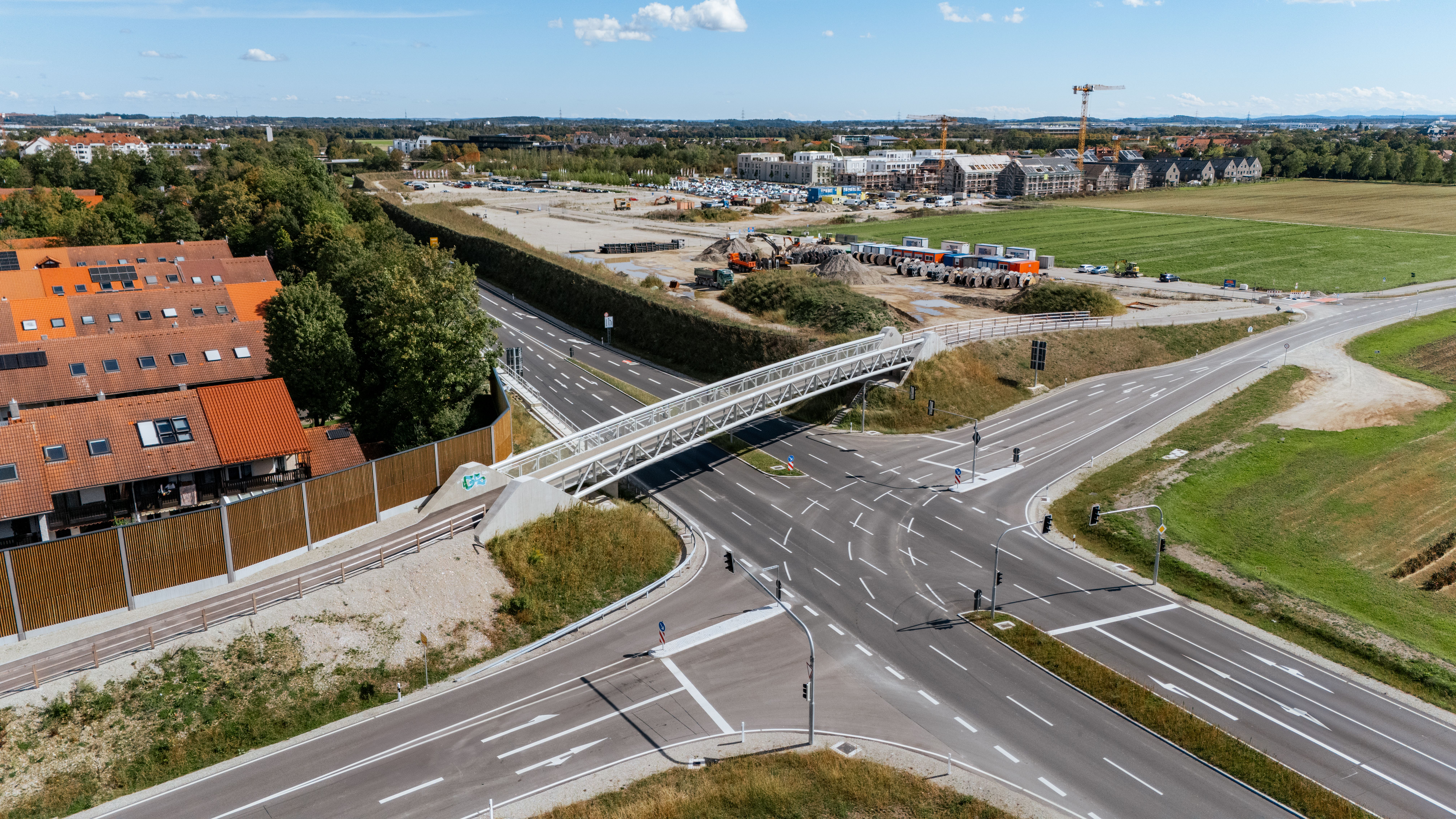 Drohnenaufnahme mit einem Gesamtblick auf die Baustelle. Zu sehen ist der komplette neue Kreuzungsbereich, darüber die neue Fußgänger- und Radbrücke sowie ein Teil der neuen Lärmschutzwand. Im Hintergrund das Baulager mit einigen JR Baufahrzeugen.